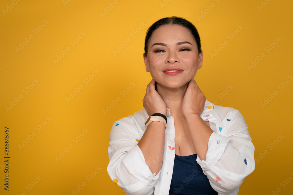 Skin care concept. Pretty woman smiles touching her neck with hands. Copy space at left side. Happy mid aged Asian woman posing in studio on yellow background. Skin care concept. Pretty woman smiles touching her neck with hands. Copy space at left side. Happy mid aged Asian woman posing in studio on yellow background.