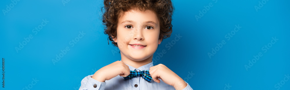 horizontal crop of curly kid in shirt touching bow tie and smiling on blue - obrazy, fototapety, plakaty 