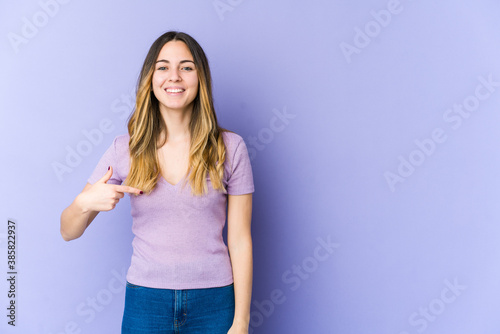 Young caucasian woman isolated on purple background person pointing by hand to a shirt copy space, proud and confident