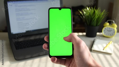 Young man holds a mobile smartphone in his hand and flips swipes right his finger on the screen on the background of the desktop, laptop