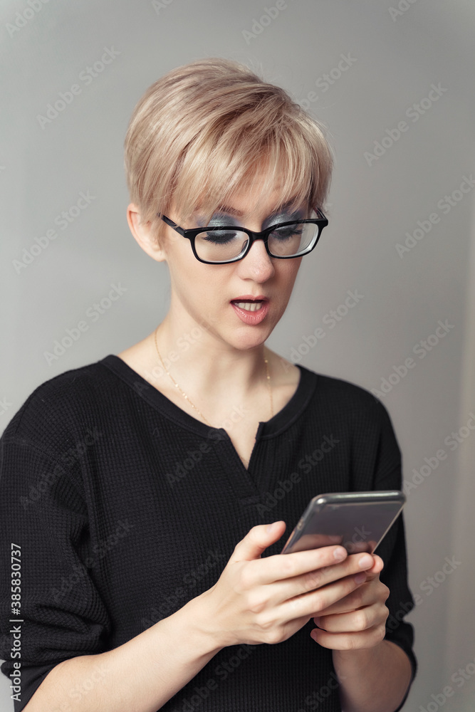 beautiful young blonde woman in glasses looks emotionally at the smartphone.