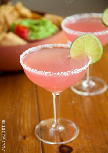 Pink Prickly Pear Margarita with Salt Rimmed Stemmed Glass with Lime Wedge on Wood Table with Chips, Salsa and Guacamole