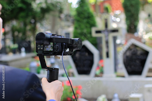 Elegant man in suit is recording grave with cross full of flowers. Funeral live streaming on cementary. Cameraman handing a microphone and camera on a gimbal.