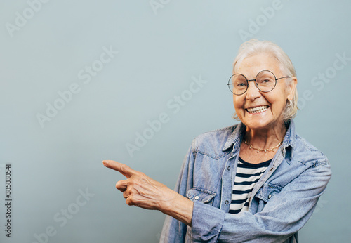 Nice friendly old woman with white hair and round glasses wants to show something to you. Stylish old ages concept. Isolated over blue background. Jeans jacket and striped shirt. photo
