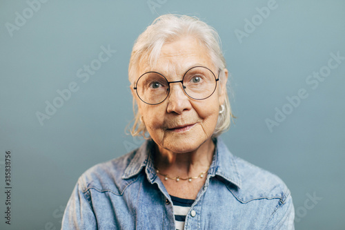 Wise concentrated old woman with attentive gray eyes in big round glasses looks into camera. Intelligent elderly lady wearing striped shirt and denim jacket isolated over gradient blue background.