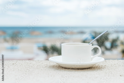 A white cup with a spoon on a saucer stands against the background of a blurred sea beach. Morning at a beautiful hotel by the sea.
