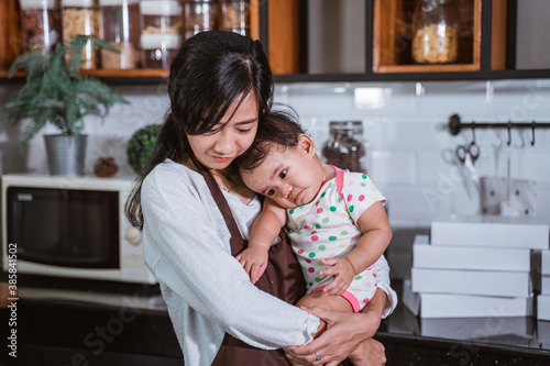 The young mother wears an apron and holds her sad baby girl while making donuts together in the kitchen