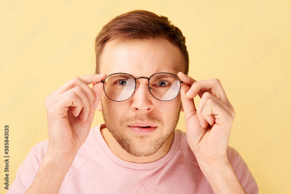 Portrait of caucasian man keeps hands on rim of spectacles, funny reaction to shocking news, gaze at the camera isolated on yellow background in casual clothes Portrait of caucasian man keeps hands on rim of spectacles, funny reaction to shocking news, gaze at the camera isolated on yellow background in casual clothes