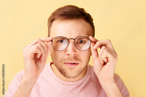 Portrait of caucasian man keeps hands on rim of spectacles  funny reaction to shocking news  gaze at the camera isolated on yellow background in casual clothes