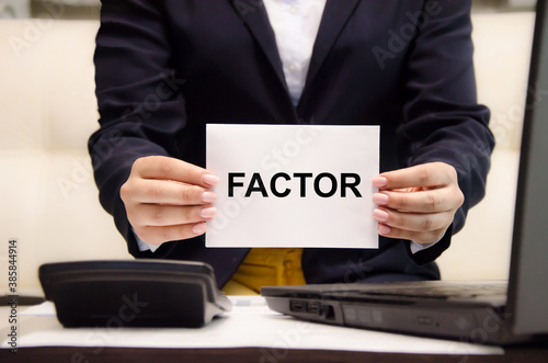 The word factor is written on a white piece of paper which is being held by a business woman. the word factor is used in business, education, medicine