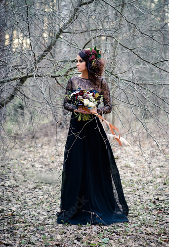 A Gothic woman in a black dress with a bouquet of flowers in a mysterious forest. Halloween concept.