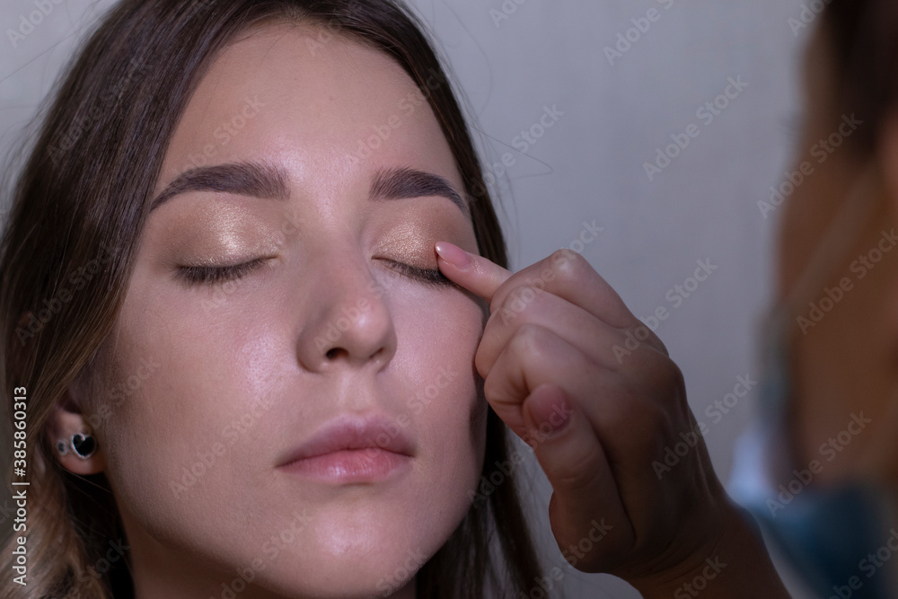 the process of applying makeup to a beautiful brunette by a master in a medical mask. beauty, modeling and cosmetics concept. the process of applying makeup to a beautiful brunette by a master in a medical mask. beauty, modeling and cosmetics concept.