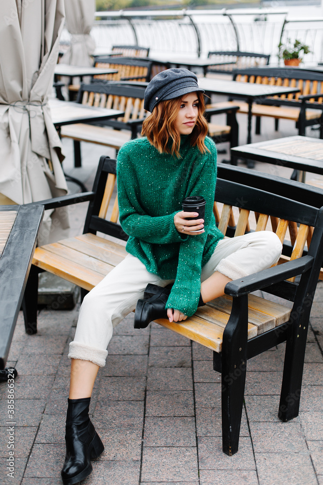 Portrait of a beautiful red-haired girl.  Beautiful red-haired girl in a green sweater and cap sits with a cup of coffee in a cafe on the street.