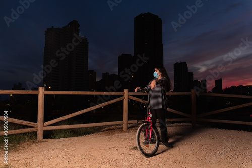 Teenager with long hair or girl looks at the city sunset with protective face mask.