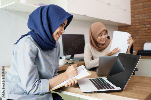 attractive cheerful young muslim business woman working on laptop and smiling while sitting at her desk modern office with her friend at studio