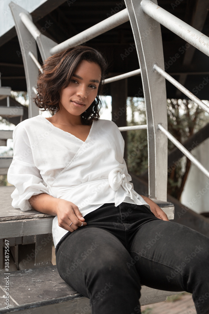 Latin young brunette woman with curls and short hair sitting relaxed on stairs, wearing a white blouse and black pants, photo in the day Latin young brunette woman with curls and short hair sitting relaxed on stairs, wearing a white blouse and black pants, photo in the day