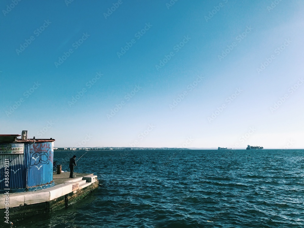 Man fishing on the dock of a bay with two ships in the distance. Blue sky, blue sea of Thessaloniki, Greece.