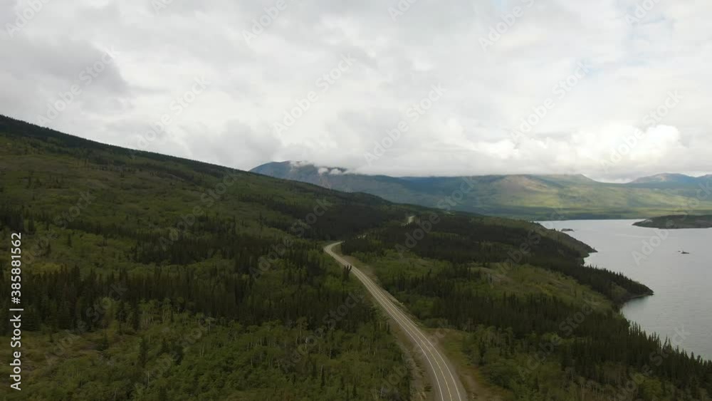 Beautiful View of Road alongside Scenic Lake surrounded by Mountains and Trees on a Cloudy Day. Aerial Drone Shot. Taken near Klondike Highway, Southern Yukon, Canada. Beautiful View of Road alongside Scenic Lake surrounded by Mountains and Trees on a Cloudy Day. Aerial Drone Shot. Taken near Klondike Highway, Southern Yukon, Canada.