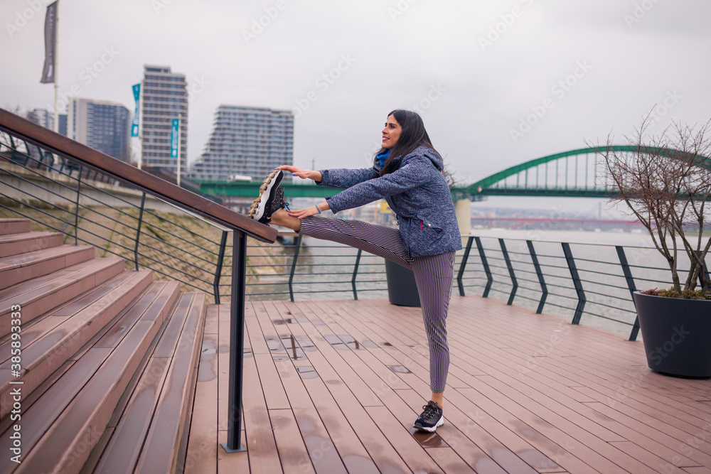 Young woman stretching legs before run. Cloudy day near river.