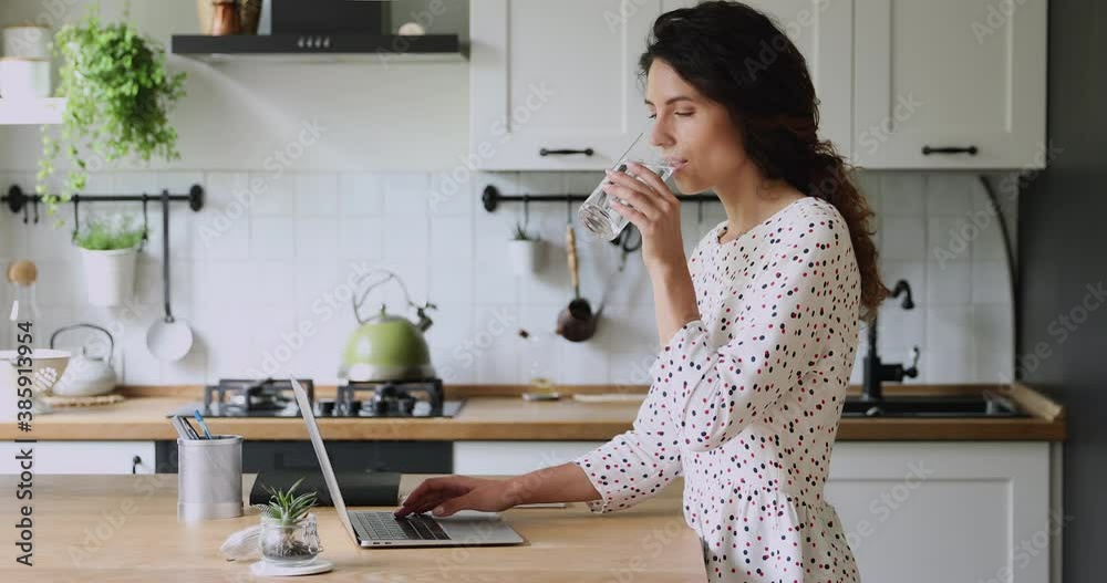 Woman standing in kitchen working on laptop do freelance remote job texting message take break drink natural mineral water makes sip caring about health, prevents dehydration. Everyday routine concept