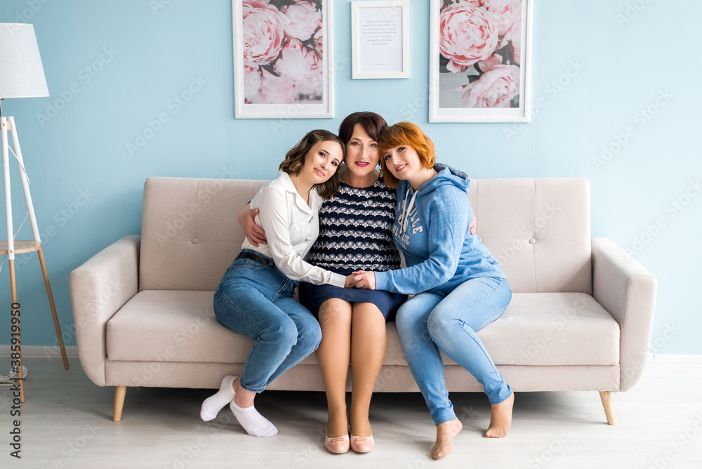Granny, her daughter and granddaughter are looking at camera and smiling while sitting on sofa at home