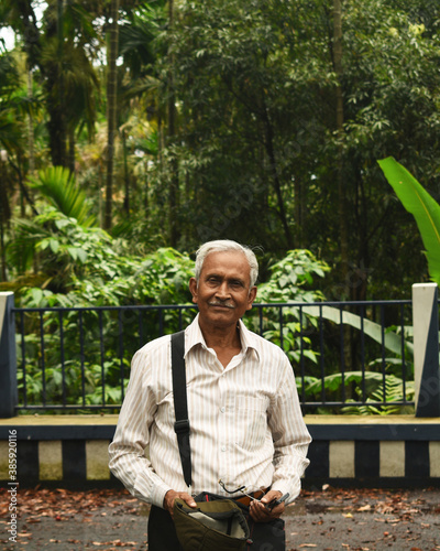 An old man, wearing spectacles and shirt, standing alone in Mawlynnong village, East Khasi Hills, Meghalaya, India. The elder person with cap in hand, side bag on shoulder, looking towards the camera.