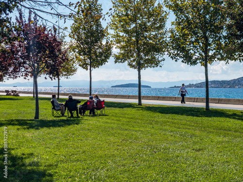 Istanbul / Turkey - 10.1.2020  New normal: A green park with trees and sea view on a sunny day. A family sits together keeping distance and wearing medical face masks to protect against Covid-19 © Stockwars
