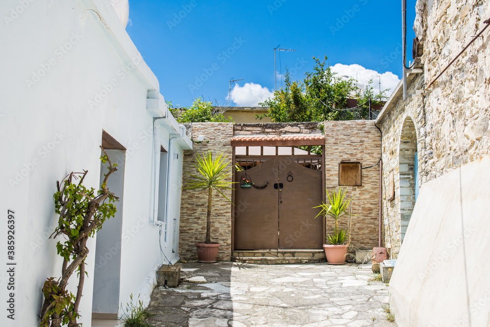 Stony alleway and entratnce to the house in Lefkara, Cyprus. Gates decorated with plants in pots over the background of blue sky. Welcome plate on the wall
