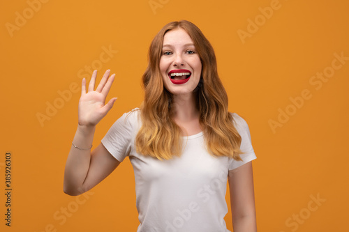 Portrait of friendly woman with wavy redhead, standing waving hand, looking at camera with engaging toothy smile