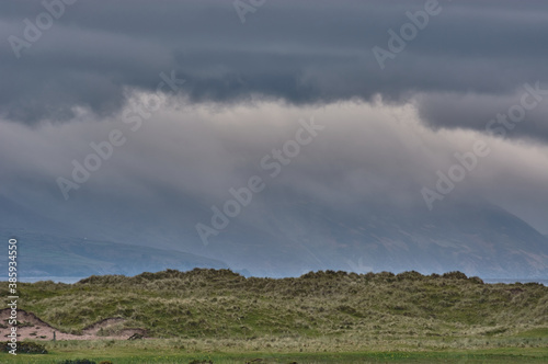 Beautiful panoramic landscape on Dingle Peninsula, Ireland. Panoramic dune landscape with mountain silhouettes and dark rain clouds in the background. Stormy weather at Inch beach in Ireland.