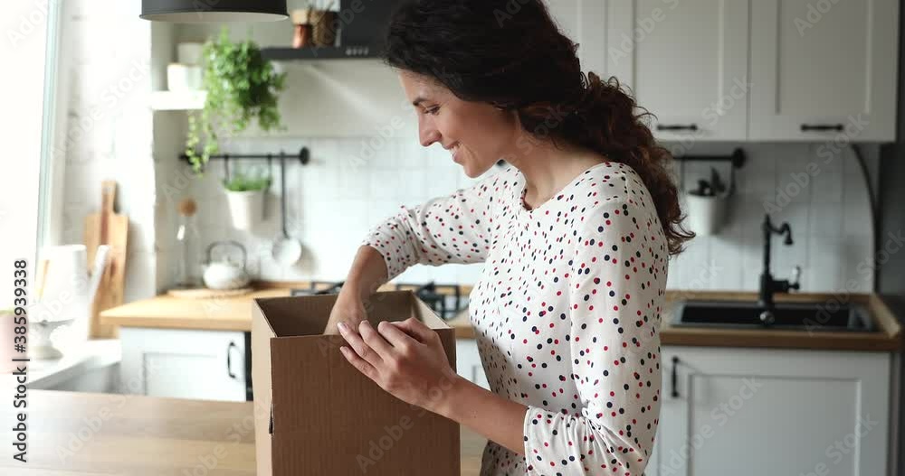 Woman received gift from foreign friend by mail, stands in kitchen opens parcel box looks inside examine carefully ordered high quality goods feels glad. Safe secure express delivery services concept Woman received gift from foreign friend by mail, stands in kitchen opens parcel box looks inside examine carefully ordered high quality goods feels glad. Safe secure express delivery services concept