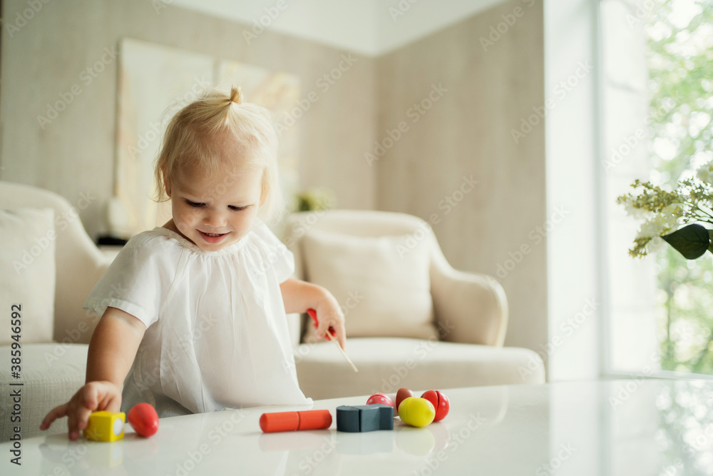 baby kids cute little blonde girl playing with her favorite colored children's toys for development, is at home in a bright room lit by the sun. Flowers on the table baby kids cute little blonde girl playing with her favorite colored children's toys for development, is at home in a bright room lit by the sun. Flowers on the table