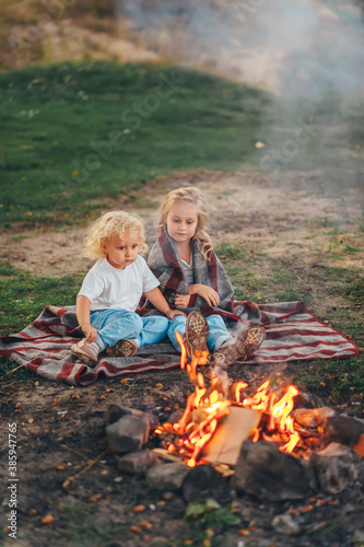 Two cute little girls sitting by a bonfire on summer evening in forest.