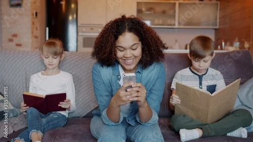 Happy mixed race nanny is using smartphone and smiling while obedient kids boy and girl are reading books at home. Babysitting and education concept.