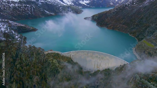 Hydropower generated from arch dam and reservoir lake in snow covered Swiss Alps mountains to produce renewable energy, sustainable hydroelectricity, aerial drone footage 4K video, autumn weather