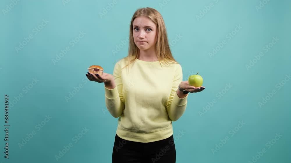 Young woman choosing between apple and cupcake, the girl is confused in her choice, isolated on a turquoise background. Young woman choosing between apple and cupcake, the girl is confused in her choice, isolated on a turquoise background.