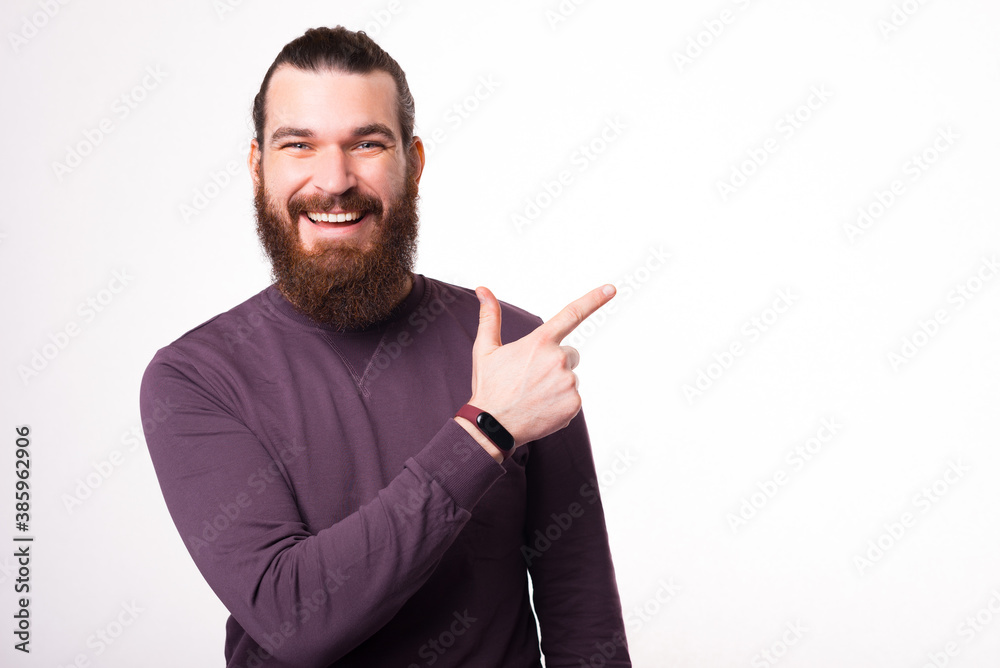 A cheerful young man is smiling at the camera and pointing with a finger away at a free space neae a white wall A cheerful young man is smiling at the camera and pointing with a finger away at a free space neae a white wall
