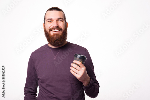 Bearded young man is smiling at the camera and holding a cup wit hot drink