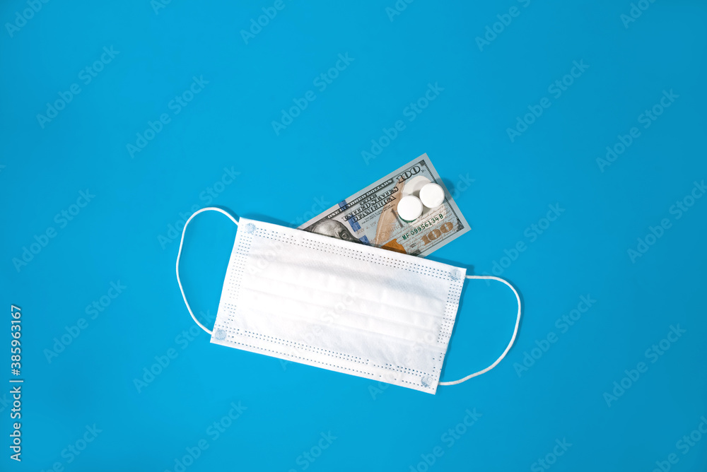 Medical face mask and tablets lie on one hundred dollar bill, blue background. Concept of expensive drugs and medicine, treatment, vaccines. Top View Medical face mask and tablets lie on one hundred dollar bill, blue background. Concept of expensive drugs and medicine, treatment, vaccines. Top View