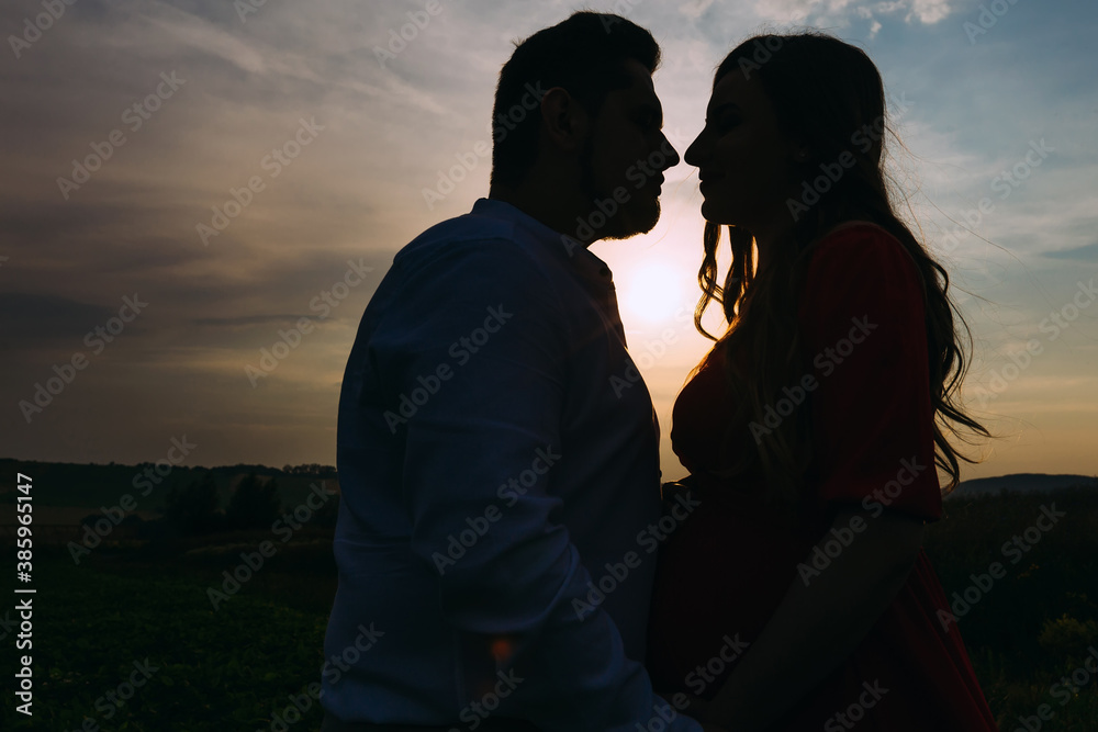 . Pregnant woman with her husband tenderly hug and kiss on the field. Couple enjoying nature and sunset