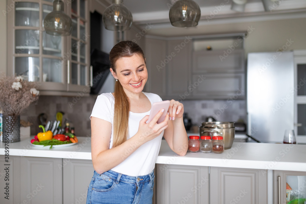 Nice young woman holds mobile phone and calls up friend, discusses recipes and smiles, standing near table on which lie fruits and vegetables. Nice young woman holds mobile phone and calls up friend, discusses recipes and smiles, standing near table on which lie fruits and vegetables.