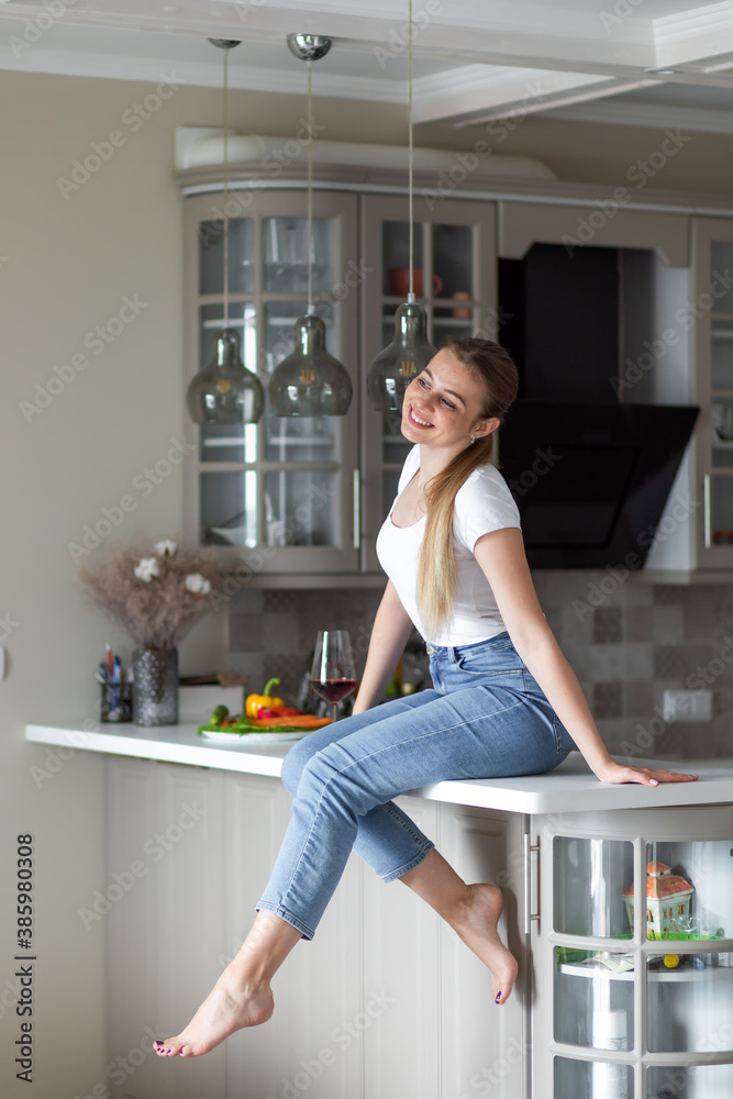 Cheerful woman sits on countertop in white modern kitchen. Morni Cheerful woman sits on countertop in white modern kitchen. Morni