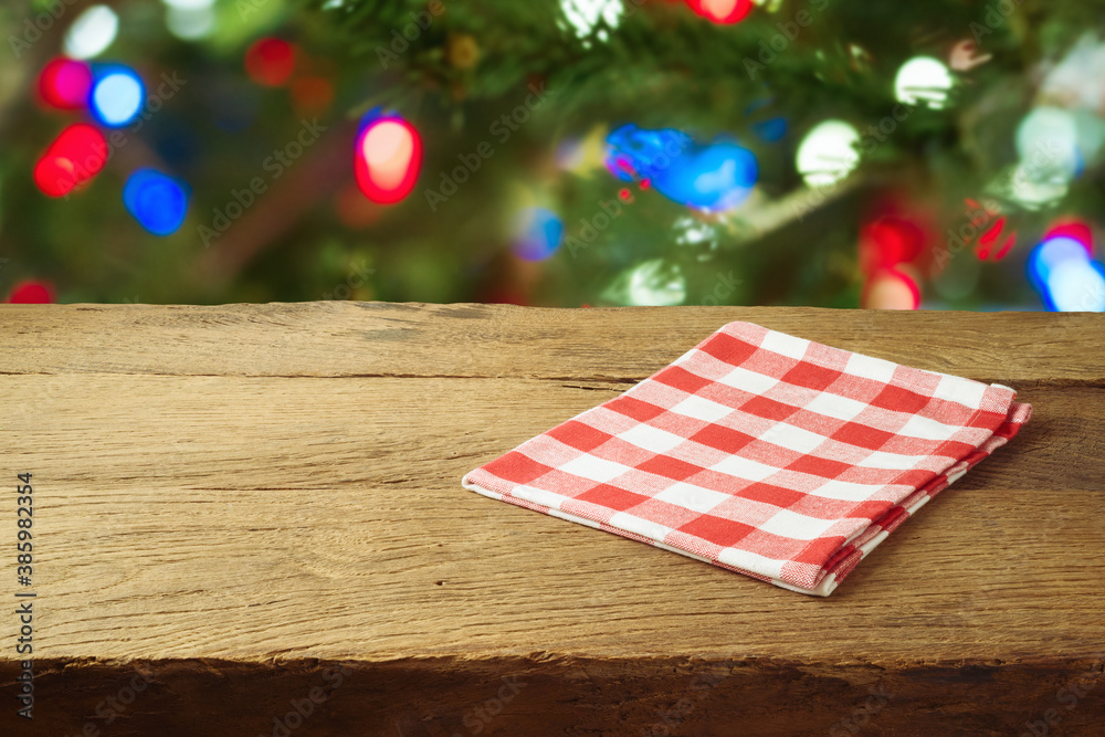 Obraz premium Christmas holiday background with empty wooden table with red checked tablecloth over Christmas tree bokeh. Product montage display or mock up design