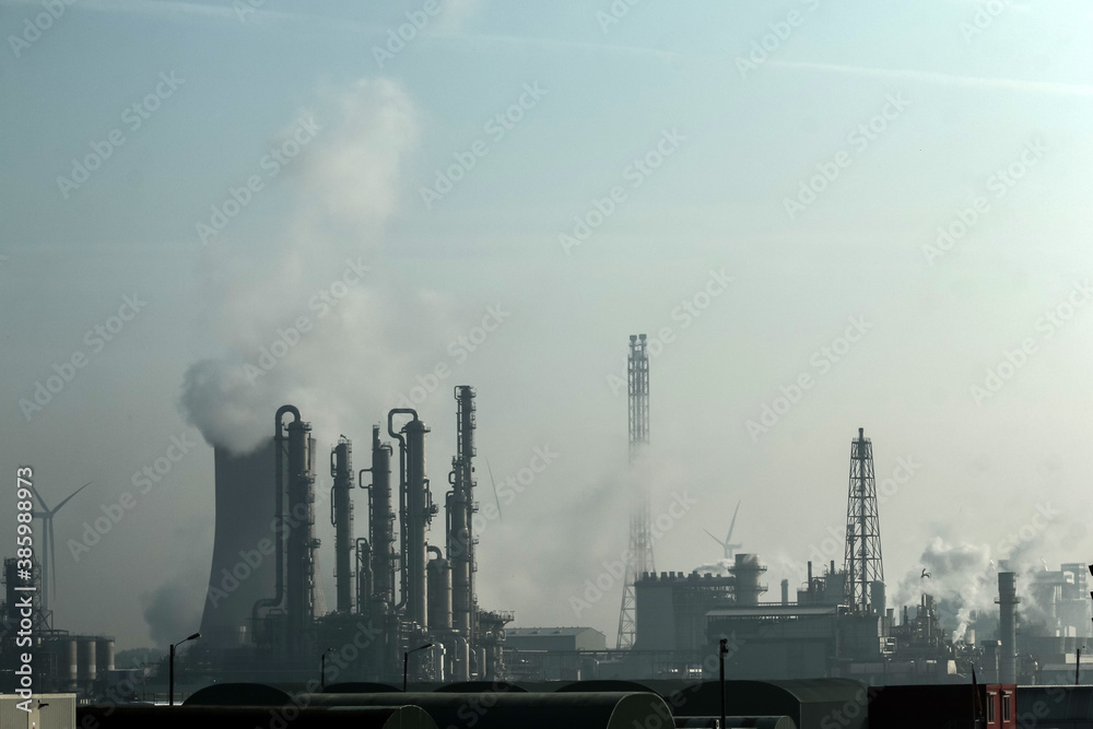 A factory with an horizon full of chimneys releasing steam or smoke. Some windmills are also visible. Symbol for environmental pollution. 