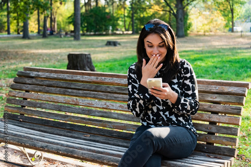Young beautiful woman sits on a bench in the park and smiles looking into a smartphone. Autumn Park