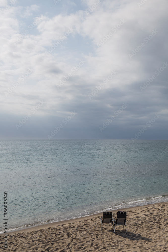 landscape of a beach, there are two chairs on the deserted shore, the sea is calm and the sky cloudy