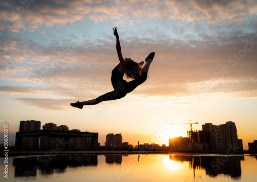 Female flexible dancer jumping during sunset on cityscape background with reflection in the water. Concept of freedom and happiness  © Hladchenko Viktor