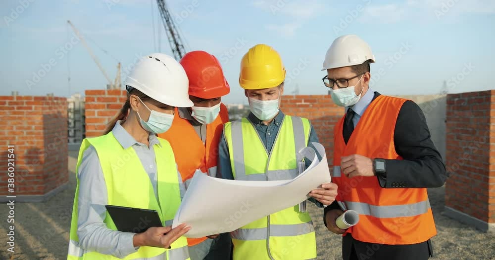 Team of mixed-races men and woman constructors in helmets and medical masks standing at building top and discussing constructing plan draft. Coronavirus concept. Engineers and architectors. Team of mixed-races men and woman constructors in helmets and medical masks standing at building top and discussing constructing plan draft. Coronavirus concept. Engineers and architectors.