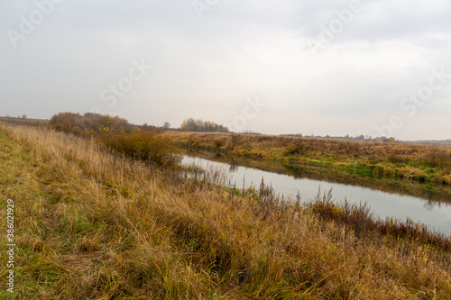 autumn rural natural landscape with calm river water reflecting yellow trees and dry grass in cloudy day