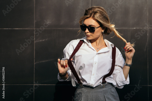 Portrait of young lady leaning on the wall of office building with clutch bag, dressed in white shirt, trousers, suspenders, looking at the camera. Image with copy space. 